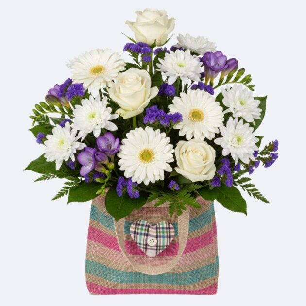 A vibrant arrangement of white roses, gerberas, and purple flowers in gift bag with a decorative heart.