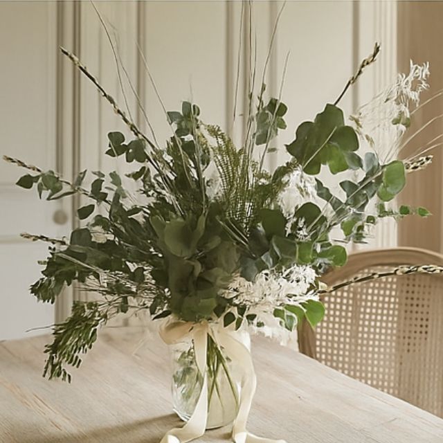 A lush, airy bouquet of assorted green and dried white foliage, eucalyptus, and tall grasses in a glass vase tied with a ribbon. Christmas foliage bouquet for sales